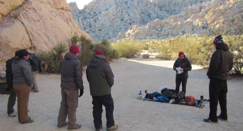 A group of people stand in the shade of large rock wall and appear to be listening to an instructor give a lesson on backpacking. There is another large rock wall in the background. 